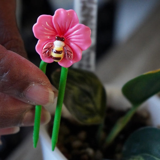 Close-up of Planty Joys decorative stem support featuring a pink flower with a bee design, held beside a small indoor plant.