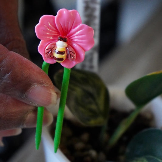 Pink flower and bee decorative stem support from Planty Joys positioned in a white pot with a small variegated indoor plant.